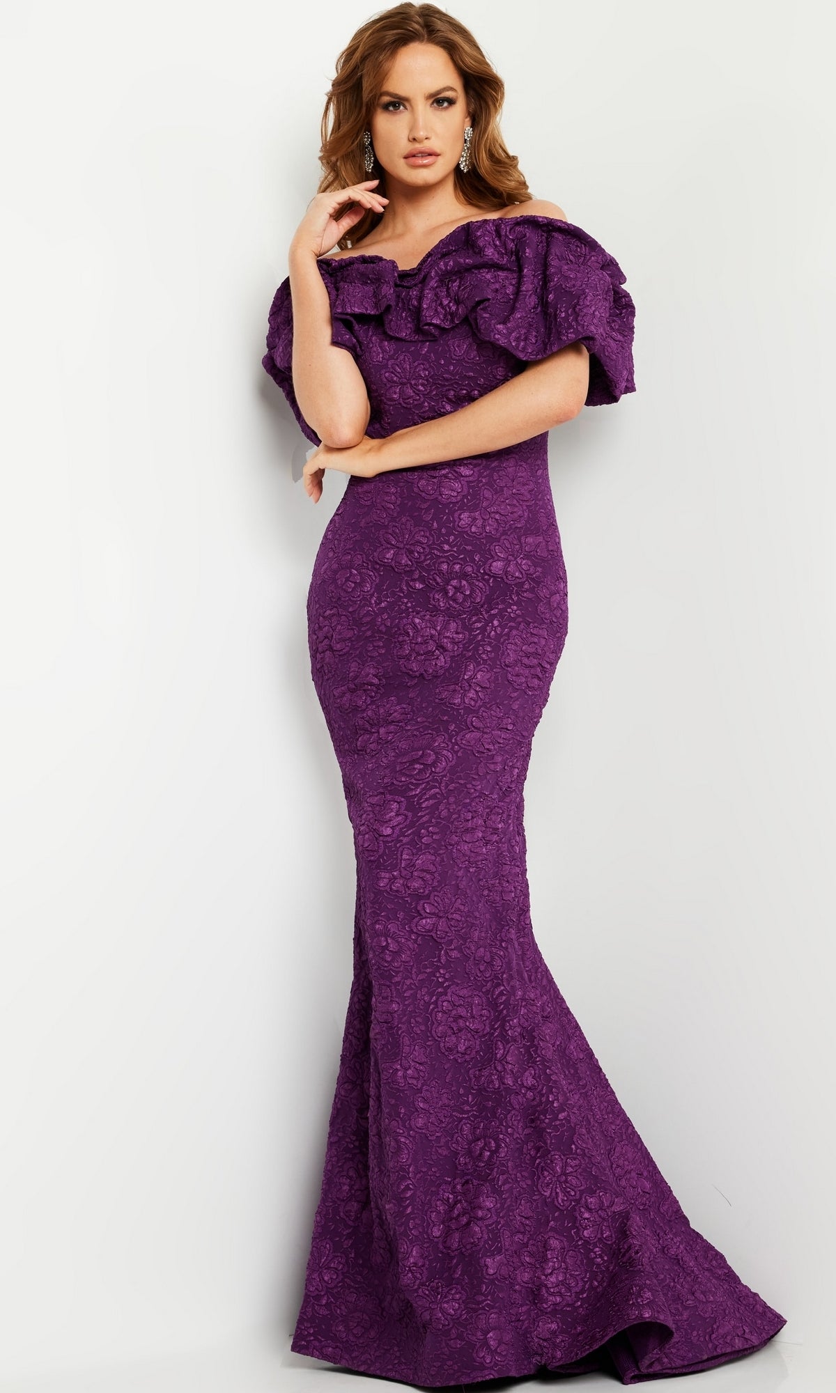 A woman stands against a light wall wearing the Jovani Long Brocade Mermaid Prom Dress 23847—a floor-length, form-fitting purple gown with ruffled off-the-shoulder sleeves.